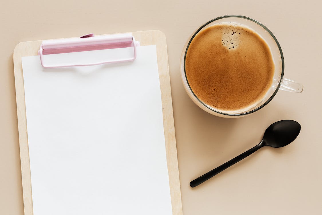 A clipboard next to a cup of coffee. A spoon sits beside the cup.