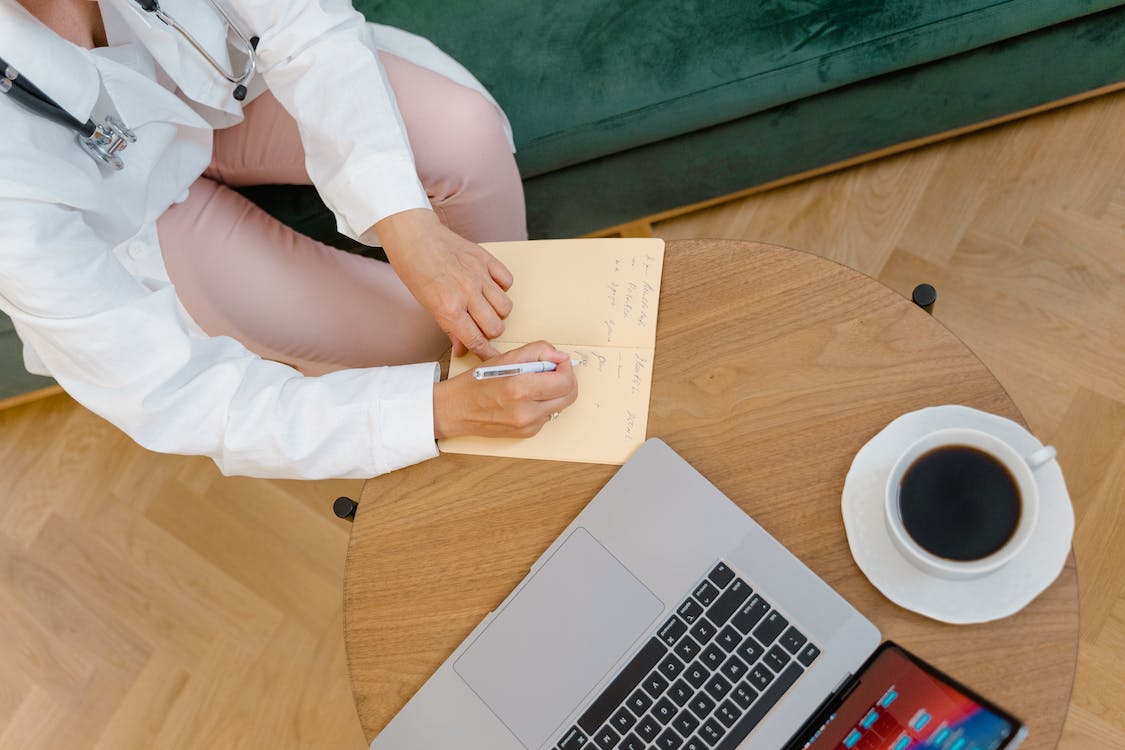 A doctor writes in a notepad on a small table with a cup of coffee and a laptop placed on top.