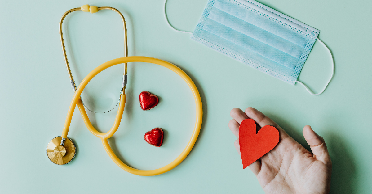 A stethoscope, disposable face mask, sit beside an open hand with a red heart in it.